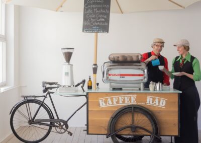 Zubereitung von Cappuccino durch einen Barista an der Espressomaschine Faema e61 mit Kaffeemühle auf dem KaffeeRad, darüber ein Sonnenschirm.