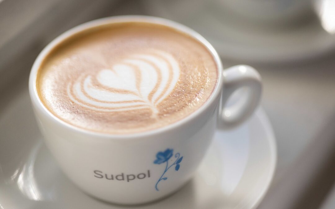 Eine Cappuccino Tasse mit einem hübschen Latte Art Herz von Sudpol in der Sonne. Die Sudpol- Blume ist auf der Tasse zu erkennen.