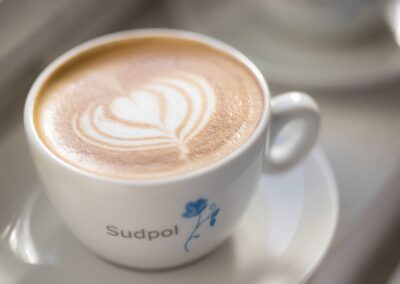 Eine Cappuccino Tasse mit einem hübschen Latte Art Herz von Sudpol in der Sonne. Die Sudpol- Blume ist auf der Tasse zu erkennen.