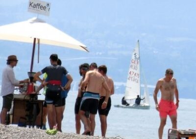 Die mobile Kaffeebar, das Sudpol KaffeeRad steht am Bodensee- Strand, ein Barista bereitet Kaffee und Espresso- Spezialitäten zu, Menschen in der Badehose stehen in der Schlange. Ein Segelboot dahinter.